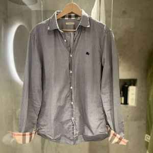 Burrberry skjorta  - Snygg grå skjorta från Burberry med klassiska rutiga detaljer på insidan av ärmsluten. Skjortan har långa ärmar och knappar framtill. Perfekt för en stilren look och är 10/10 i skick knappt använd säljer till ett riktigt bra pris för passar ej mig skjortan pssar mellan M-L