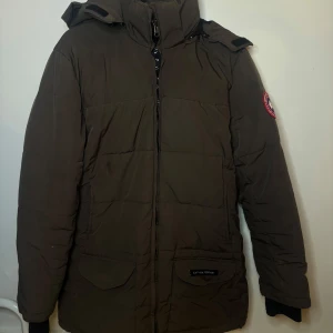 Brun parkajacka från Canada Goose - Säljer en brun parkajacka från Canada Goose med huva och pälsdetalj. Jackan är äkta men köpt för några år sedan därav inget kvitto. Säljer den billigt därför. Päls finns men dragkedjan behöver fixas på luvan. 