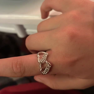 Silverfärgad ring med hjärtdetaljer - Elegant silverfärgad ring med två hjärtformade detaljer prydda med glittrande stenar. Perfekt för att ge en romantisk touch till din stil.