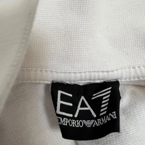Vit tröja från Emporio Armani EA7 - Snygg vit kofta från Emporio Armani EA7 med stilren design. Tröjan har en diskret logga på bröstet och ett randigt mönster på baksidan nära nacken. Perfekt för en minimalistisk look.