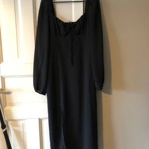 Svart klänning från Gina Tricot - Elegant svart klänning från Gina Tricot med långa ärmar och knytning framtill. Perfekt för en stilren look med en subtil slits för extra rörelsefrihet. ALDRIG ANVÄND