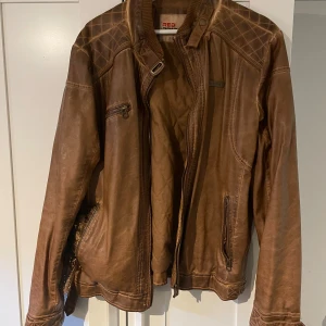 Brun skinnjacka från Red - Snygg brun skinnjacka från Red med quiltade detaljer på axlarna. Jackan har dragkedja framtill och flera praktiska fickor. Tillverkad i polyuretan med polyesterfoder för extra komfort. Perfekt för en cool och avslappnad stil.