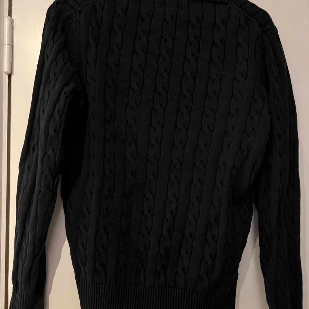 Snygg svart kabelstickad tröja från GANT i premium bomull. Tröjan har en klassisk rund halsringning och ribbade muddar vid ärmslut och nederkant. Perfekt för en stilren look.. Neuletakit & Villapaidat.