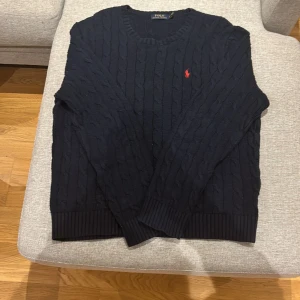 Mörkblå stickad tröja från Ralph Lauren - Säljer en stilren mörkblå stickad tröja från Ralph Lauren med det klassiska röda logotypbroderiet på bröstet. Tröjan har en kabelstickad design och långa ärmar, perfekt för kyligare dagar.