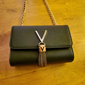 Valentino väska  - Elegant svart axelväska med guldkedja och dekorativt V-emblem. Väskan har en stilren design med en kedja som ger en lyxig känsla. Perfekt för att lyfta vilken outfit som helst.