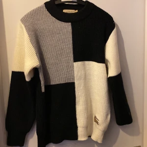 Svart och vit stickad tröja från burberry - Snygg stickad tröja från Burberry i svart, vit och grå blockdesign. Tröjan har en rund hals och långa ärmar, perfekt för kyligare dagar. Den unika färgblocksstilen ger en modern touch.