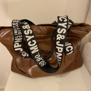 Brun axelväska med tryckta band - Snygg brun axelväska i syntetmaterial med breda svarta axelband som har vit text. Väskan har en praktisk dragkedja upptill och en rymlig design, perfekt för vardagsbruk.