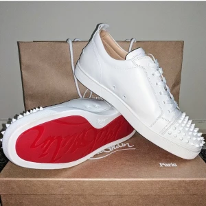 Vita loubs - Stilrena vita sneakers med nitar på tån och röd sula. Skorna har snörning och är tillverkade i skinn, vilket ger en lyxig känsla. Perfekta för att ge en edgy touch till din outfit.