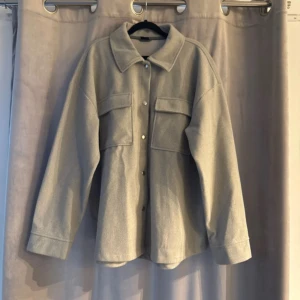 Grå overshirt från Gina Tricot - Snygg grå overshirt från Gina Tricot med knappar framtill och två stora bröstfickor. Perfekt för lager-på-lager-stil. Den har en klassisk krage och långa ärmar.