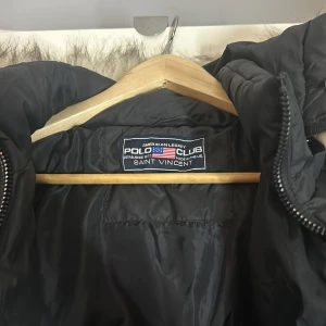 Svart pufferjacka från Polo Club - Snygg svart pufferjacka från Polo Club med fuskpäls på huvan. Jackan har en dragkedja framtill och en logotypdetalj på ärmen. Perfekt för kyliga dagar.