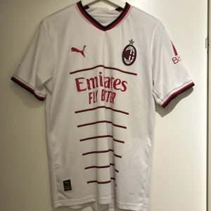 Vit AC Milan fotbollströja från Puma - Snygg vit AC Milan fotbollströja från Puma med röda och svarta detaljer. Tröjan har korta ärmar och klubbens emblem på bröstet. Perfekt för fans som vill visa sitt stöd med stil! ⚽️