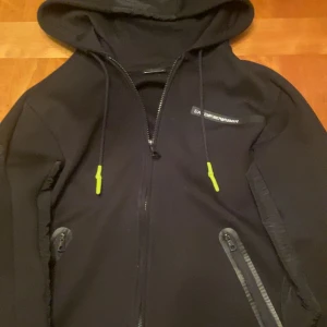 Svart hoodie från Emporio Armani - Snygg svart hoodie från Emporio Armani med dragkedja och huva. Den har stilrena detaljer och logotyp på bröstet. Ända felet är att högra fickans dragkedja är lite krånglig. Använd fåtal gånger.