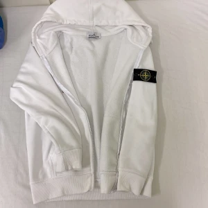 Vit hoodie från Stone Island - Säljer en stilren vit hoodie från Stone Island med dragkedja och klassisk logga på ärmen. Den har en bekväm passform och är tillverkad i mjukt material. Inga skador i flickar. Tröjan är på bra skick. Priset diskuteras på en snabb affär