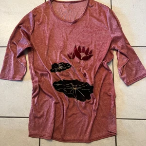 Röd topp med blommönster - Säljer en röd topp med ett unikt blommönster i svart och rött framtill. Toppen har trekvartsärmar och en rund halsringning. Perfekt för en avslappnad stil.