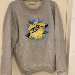 Grå sweatshirt från Maison Kitsuné - Säljer en grå sweatshirt från Maison Kitsuné med ett färgglatt citronmotiv på framsidan. Tröjan har rund hals och långa ärmar, perfekt för en avslappnad stil