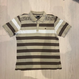 DC piké - Snygg brun- och vitrandig pikétröja från DC med krage och knappar. Den har korta ärmar och ett diskret mönster på axlarna samt broderad logga på bröstet. Riktigt fet. Har ett fåtal små och näst intill osynliga hål då den är begagnad.