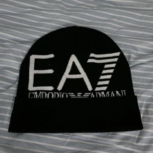 Svart mössa från Emporio Armani Ea7 - Snygg och stilren mössa från Emporio Armani i svart och grått med stor EA7-logga. Perfekt för att hålla värmen med stil under kyliga dagar. Knappt använd så inga tecken på användning eller slitning. 