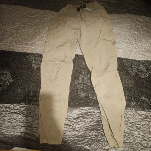 Beige Peak Performance cargopants - Säljer ett par beige cargopants med flera fickor och elastiska muddar vid bensluten. Perfekta för en avslappnad stil och praktiska med sina många fickor. Passar bra till vardagsäventyr.
