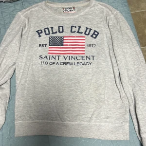 Grå tröja från Saint Vincent - Säljer en grå tröja från Saint Vincent med tryck av amerikansk flagga och texten 'Polo Club'. Tröjan har långa ärmar och rund halsringning. Perfekt för en avslappnad stil.