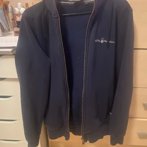 Hoodie från Sail Racing - En marinblå zip up hoodie från Sail Racing med dragkedja och broderad logga på bröstet. Jackan har en huva och långa ärmar. Väldigt bra nu för vintern. Säljer till brorsan. Köpte för 1000kr 