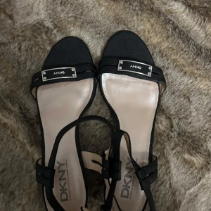 Svarta sandaler från DKNY - Stilrena svarta sandaler från DKNY med öppen tå och justerbar rem runt ankeln. Sandalerna har en metallisk detalj med logotyp på framsidan och en bekväm klack. 