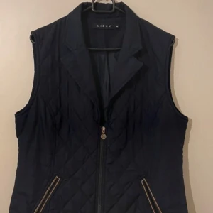 Mörkblå quiltad väst från Micha M - Snygg mörkblå quiltad väst från Micha M med dragkedja framtill och praktiska fickor. Perfekt för lager-på-lager under kyliga dagar. En stilren och mångsidig väst för alla tillfällen.