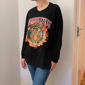 Svart långärmad tröja med tigertryck från FB Sister - Cool svart långärmad tröja från FB Sister med ett stort tigertryck och texten 'Courageous' i rött och orange. Perfekt för en avslappnad stil eller att sticka ut lite extra. Tröjan är i bra skick och passar perfekt till jeans eller leggings.