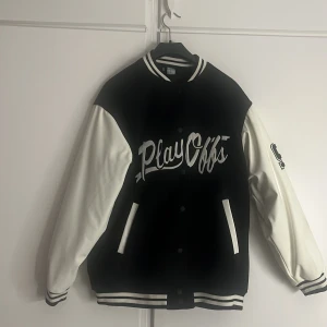 Svart och vit bomberjacka med PlayOffs-tryck - Säljer en svart och vit bomberjacka med texten 'PlayOffs' på bröstet. Jackan har långa ärmar och ett  nummer '92' på ärmen. Perfekt för höst och vår, och den har knappar framtill. Passar både till vardags och lite mer avslappnade tillfällen. Använd bara 1 gång.