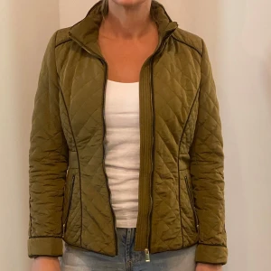 Olivgrön quiltad jacka - Säljer en olivgrön quiltad jacka i nyskick. Den har en dragkedja framtill och långa ärmar. Perfekt för höst och vår med sin stilrena design och bekväma passform. Jackan är både snygg och praktisk för kyligare dagar.
