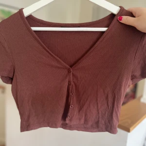 Brun ribbad croppad topp - Säljer en superfin brun ribbad topp med korta ärmar och v-ringning. Den är croppad och har knappar framtill, perfekt för sommardagar eller en avslappnad höstlook. Toppen är i bra skick och passar perfekt till jeans eller kjol!