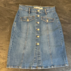 Jeanskjol med knappar - Säljer en snygg jeanskjol från Bianca i storlek S. Kjolen har en klassisk blå denimfärg och är dekorerad med knappar framtill. Den har också två fickor med lock och bälteshällor i midjan. Perfekt för en avslappnad vardagslook!