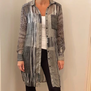 Mönstrad lång skjorta från KappAhl - Säljer en supercool lång skjorta från KappAhl i storlek 34. Den har ett unikt patchwork-mönster i svart, vitt och beige med långa ärmar och knappar framtill. Perfekt för höst och vår, och kan stylas både upp och ner beroende på tillfälle. Ett måste för garderoben! 🌟