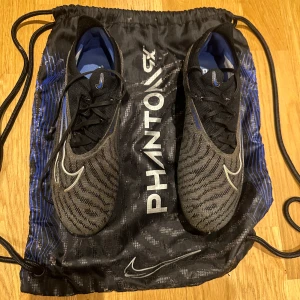Nike Phantom Gx fotbollsskor - Säljer ett par Nike Phantom Gx elite fotbollsskor i svart och blått. Skorna har en snygg design med snörning och är perfekta för fotbollsspel. De kommer med en praktisk skopåse för enkel transport.