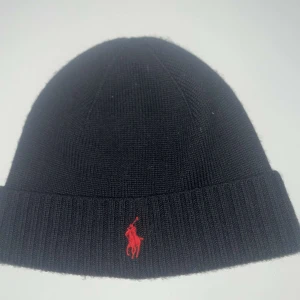Svart mössa från Polo Ralph Lauren - Snygg svart mössa från Polo Ralph Lauren i merinoull. Den har en röd broderad logotyp framtill och är perfekt för kyliga dagar. Mössan är ribbad och ger en stilren look.