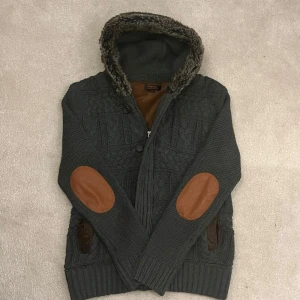 Fodrad Cardigan med Päls Huva - Säljer en mörkgrå fodrad cardigan med pälskrage och bruna armbågslappar. Jackan har knappar och dragkedja framtill samt ribbade muddar. Perfekt för kyliga dagar! Det står L i men sitter mer som M. Vid minsta fråga eller fundering hör av er🤝