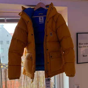 Fjällräven jacka - Säljer en snygg gul dunjacka från Fjällräven Expedition Series. Jackan har en blå insida och är utrustad med en huva och dragkedja. Perfekt för kalla dagar med sin vadderade design och praktiska fickor. Pris kan diskuteras vid snabbaffär