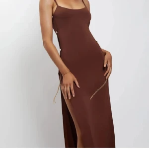 Brun långklänning med snörning - Snygg brun långklänning med smala axelband och stilren design. Klänningen har en slits på sidan och dekorativ snörning längs sidorna som ger en unik touch. Perfekt för en elegant look.