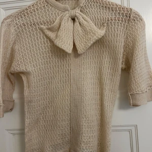 Beige stickad tröja med rosett - Säljer en  beige stickad tröja med en stor rosett framtill. Tröjan har trekvartsärmar och ett luftigt stickat mönster. Perfekt för lager-på-lager-stil.