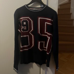 Mörkblå/marinblå tröja från Hilfiger Denim - Säljer en mörkblå långärmad tröja från Hilfiger Denim med stora siffror '85' i rött och vitt på framsidan. Tröjan har en rund halsringning med randiga detaljer. Perfekt för en avslappnad, streetwear  stil.