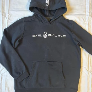 Svart hoodie från Sail Racing - Säljer en svart hoodie från Sail Racing med vit logga på bröstet och huvan. Tröjan har en klassisk känguruficka och långa ärmar. Perfekt för en avslappnad stil.