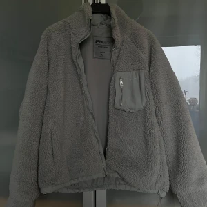 Grå teddyjacka  - Mysig grå teddyjacka från FB Sister med dragkedja och en praktisk bröstficka. Perfekt för kyliga dagar när du vill hålla dig varm och bekväm. Jackan har en stilren design med en mjuk och fluffig yta. Passar perfekt till våren som jacka.  Man kan även ha den som kofta 💞