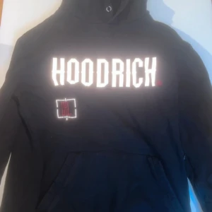 Svart hoodie från Hoodrich - Säljer en svart hoodie från Hoodrich med stor vit logga på framsidan och detaljerad design på baksidan. Hoodien har en klassisk känguruficka och justerbar huva med dragsko. Perfekt för en avslappnad stil.