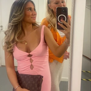 Rosa klänning med utskärningar - Snygg rosa klänning med djup v-ringning och trendiga utskärningar framtill. Perfekt för en kväll ute eller en speciell tillställning. Klänningen har en figurnära passform som framhäver silhuetten.