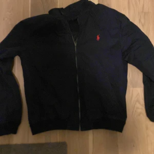 Blå  jacka från Polo Ralph Lauren - Säljer en stilren svart jacka från Polo Ralph Lauren med dragkedja och röd logga på bröstet. Jackan har en klassisk design med långärmade ärmar och ett rutigt foder i grönt och blått. Perfekt för en snygg och avslappnad look.