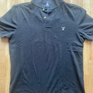 Svart pikétröja från GANT - Säljer en  svart pikétröja från GANT med korta ärmar och broderad logga på bröstet. Tröjan har en stilren design med knappar vid halsen och är perfekt för en avslappnad look. Storlek: Large Hör av dig vid några frågor.😊