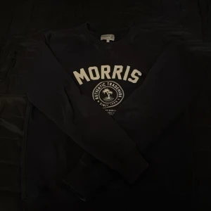 Svart tröja från Morris - Säljer en svart tröja från Morris med tryck på framsidan. Tröjan har långa ärmar och en klassisk rund halsringning. Perfekt för en stilren och avslappnad look.