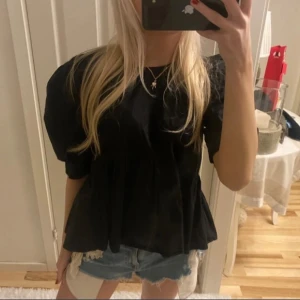 Svart blus med volangdetaljer - Snygg svart blus med volangdetaljer och korta ärmar. Perfekt för en stilren look. Blusen har en lös passform och är tillverkad i ett mjukt material. Lånade bilder från förra ägaren ❤️ storlek m men passar en xs