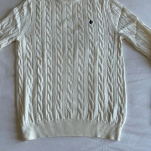 Vit Morris tröja  - Säljer en stilren vit kabelstickad tröja från Morris i 100% pima bomull. Tröjan har ett diskret broderat emblem på bröstet och ribbade muddar vid ärmslut och nederkant. Perfekt för en klassisk look!