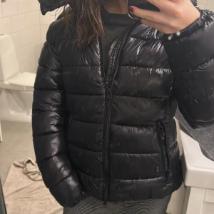 Svart pufferjacka från 157 - Säljer en svart pufferjacka från 157 i storlek M. Jackan har en glansig yta med dragkedja framtill och två praktiska fickor med dragkedjor. Perfekt för kyliga dagar med sin vadderade design. Huvan ger extra skydd mot väder och vind. HELT NY!! SITTER SOM STORLEK S