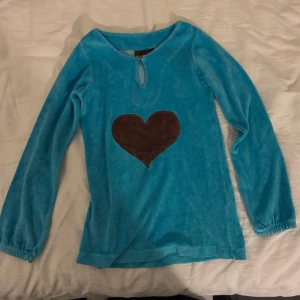 Blå långärmad klänning med hjärtmönster från Me & I - Säljer en söt blå långärmad klänning från Me & i med ett brunt hjärta på framsidan. Tröjan har en liten knappdetalj vid halsen och är tillverkad i ett mjukt material. 